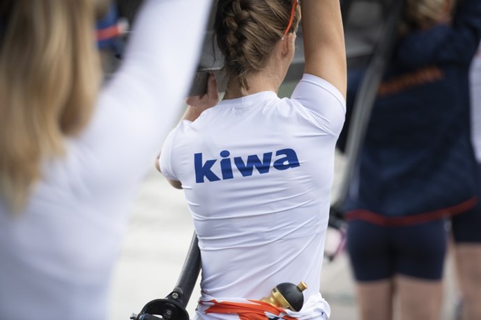 To jenter med Kiwa logo på ryggen bærer en kayakk. Bilde.