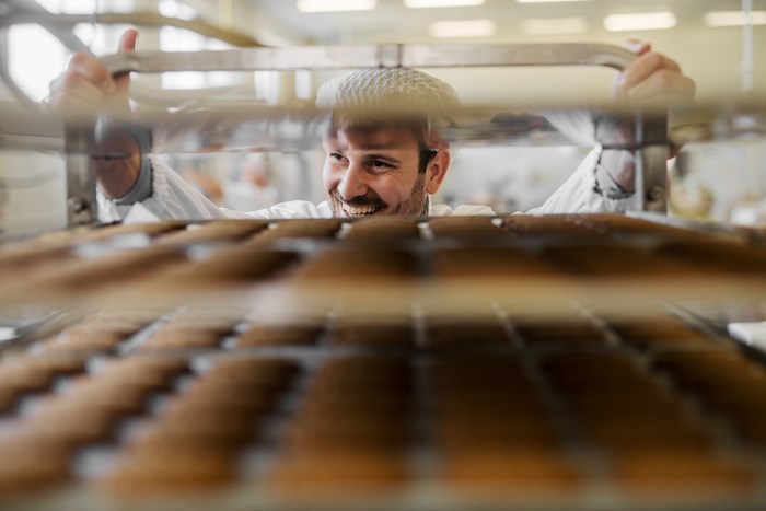 En leende man rullar en ställning med flera bakplåtar fulla med bröd på ett bageri.