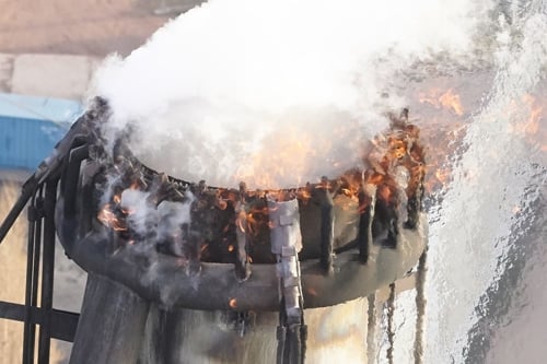 Industrial chimney with fire at the edge, emitting white smoke against a blurred background