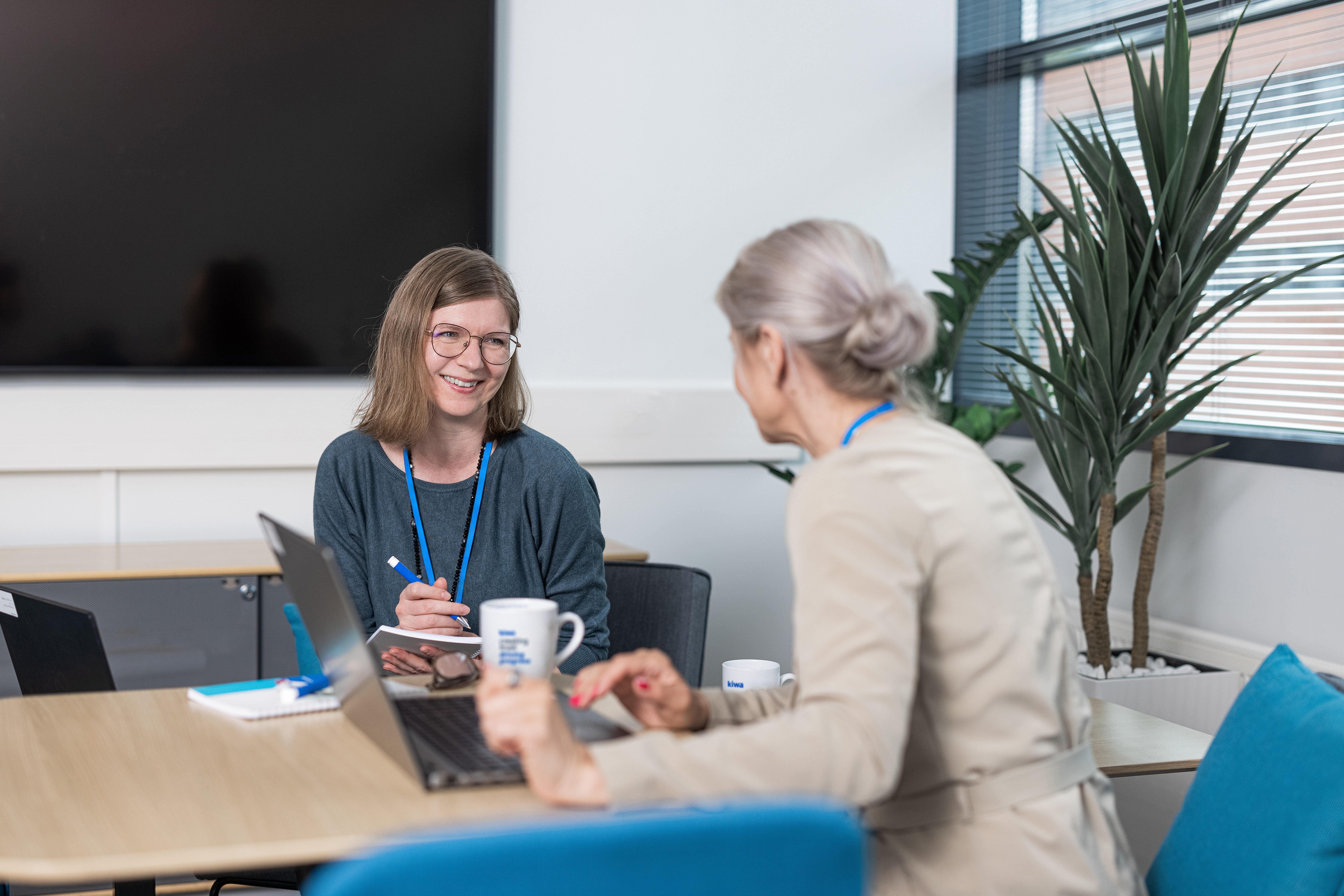 Two women discussing business over laptops in a modern office meeting room with coffee mugs and a large plant.