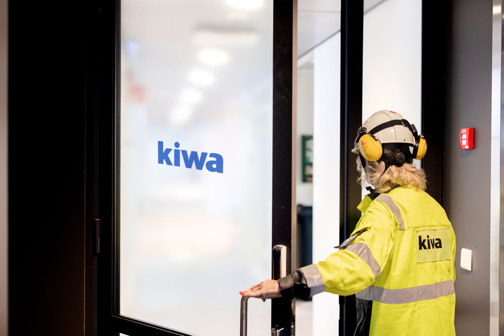 Worker in high-visibility safety gear opening a glass door with Kiwa logo, wearing hard hat and hearing protection in an office corridor.