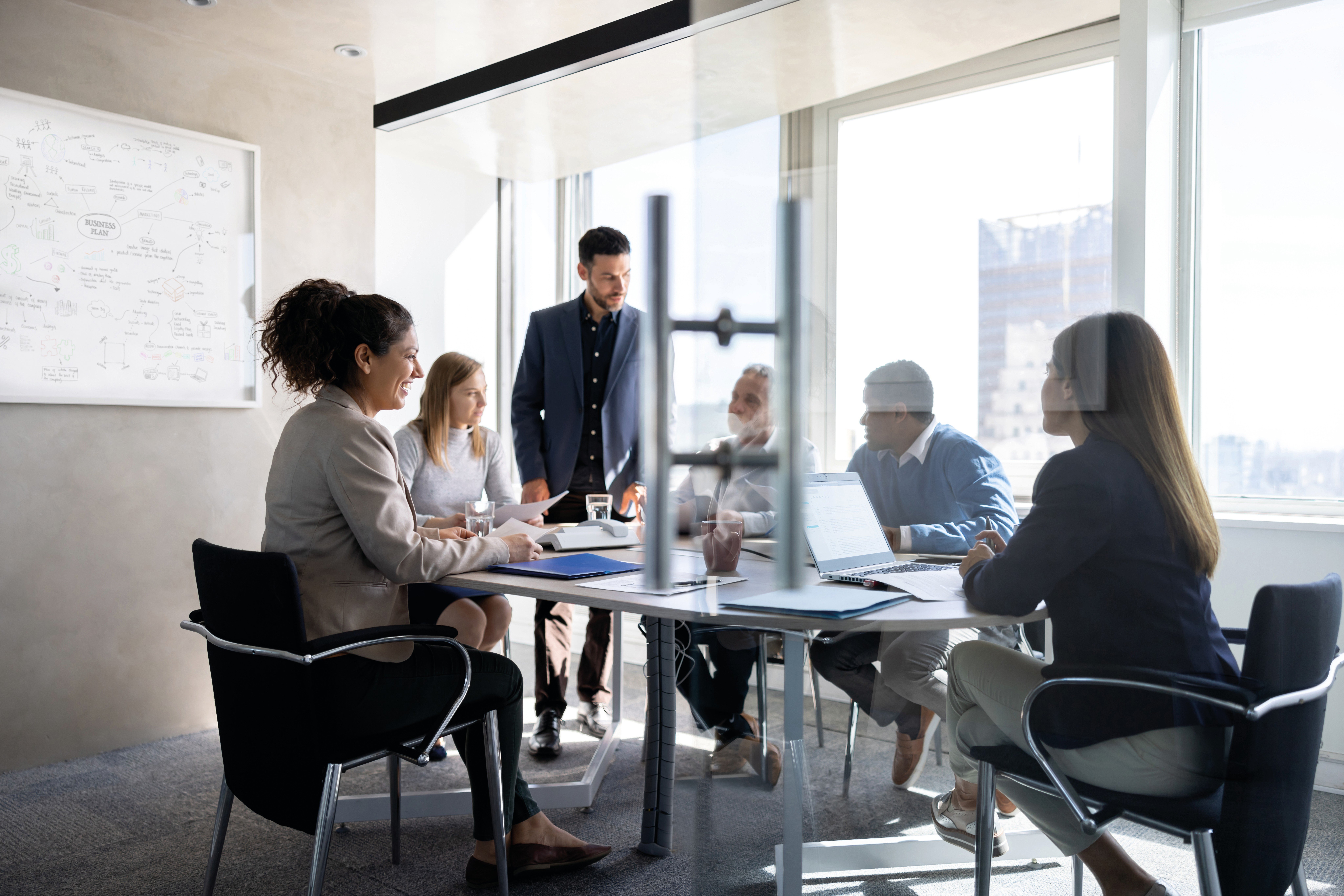Business team meeting in a modern glass conference room with laptops, documents, and a whiteboard.