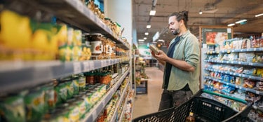 Man shopping in a grocery store aisle, reading product label and pushing a cart filled with groceries