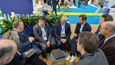 A group of business professionals in suits having a discussion at a trade show booth, surrounded by plants and a blue partition, with an exhibition floor in the background