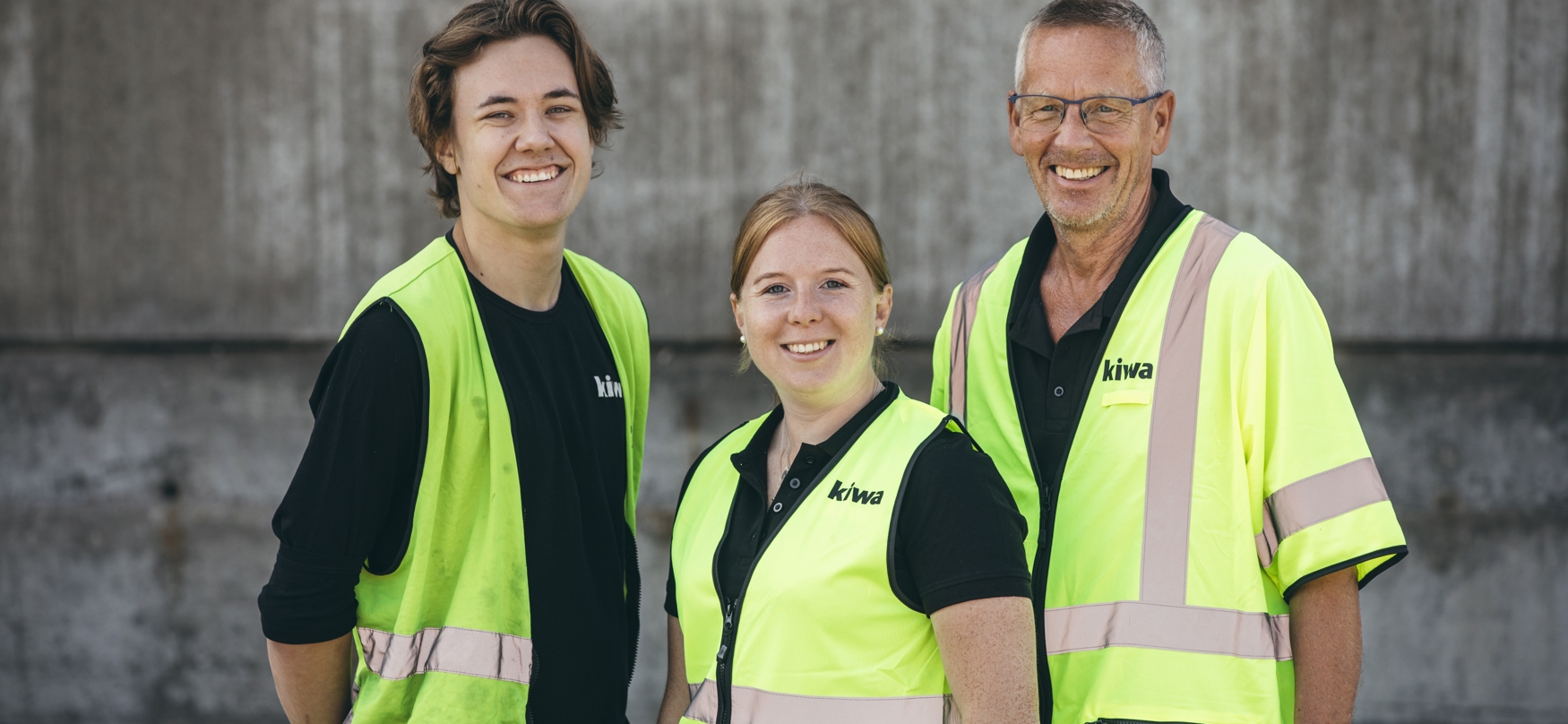 Drei Arbeiter in neongelben Sicherheitswesten und schwarzen Hemden stehen vor einer Betonmauer.