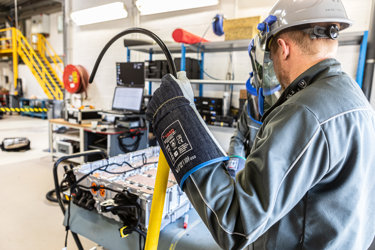 Technician wearing a safety helmet, face shield, and protective gloves working on an electric vehicle battery pack in an industrial workshop, handling insulated cables during battery maintenance or inspection.