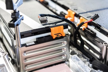 Close-up of industrial battery modules with metallic casings, orange locking mechanisms, and black electrical connectors connected by insulated wiring, showing components of an advanced battery system.