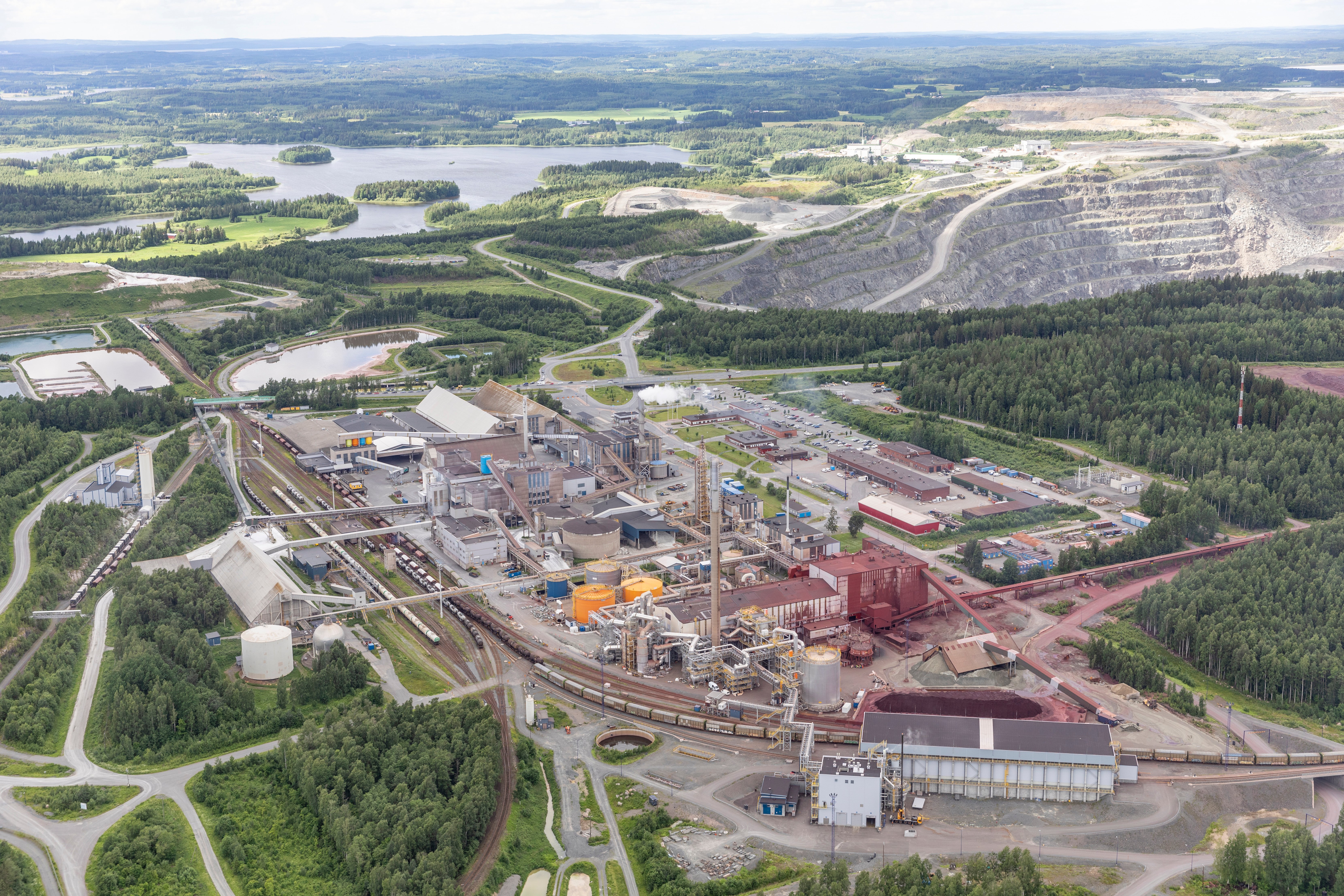 Aerial view of an industrial mining and processing plant surrounded by forest, lakes, and terraced quarry pits.