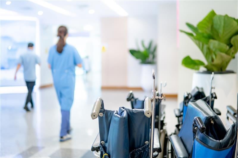 Hospital corridor with two blurred medical staff members walking past a row of wheelchairs and a potted plant.