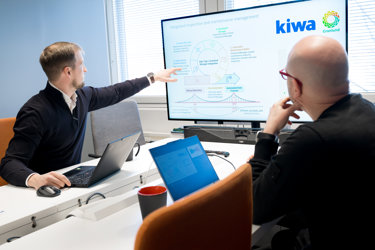 Two professionals in an office meeting discussing integrated inspection and maintenance management. One points to a large screen displaying a process diagram with Kiwa and Granlund logos, while the other listens. Laptops are open on the table.