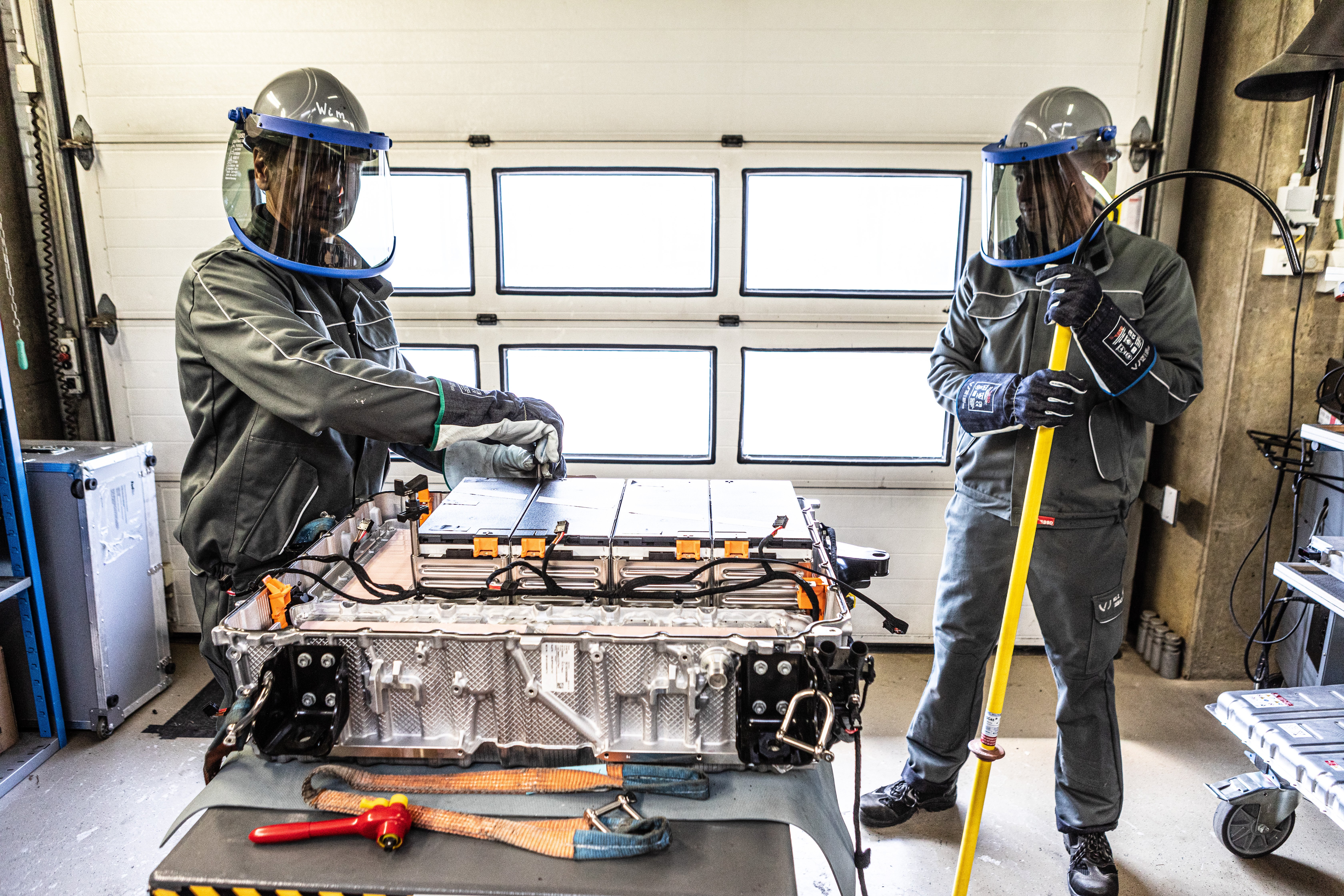 Two workers in protective gear inspect a large electric vehicle battery pack in a workshop.