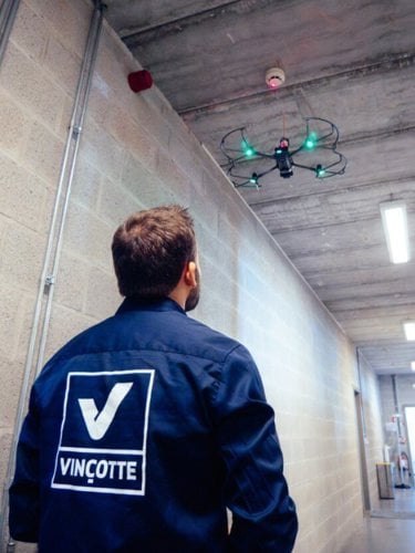 A Vincotte employee in a Vincotte jacket operates a drone with green lights in an industrial corridor, showcasing technology and inspection services