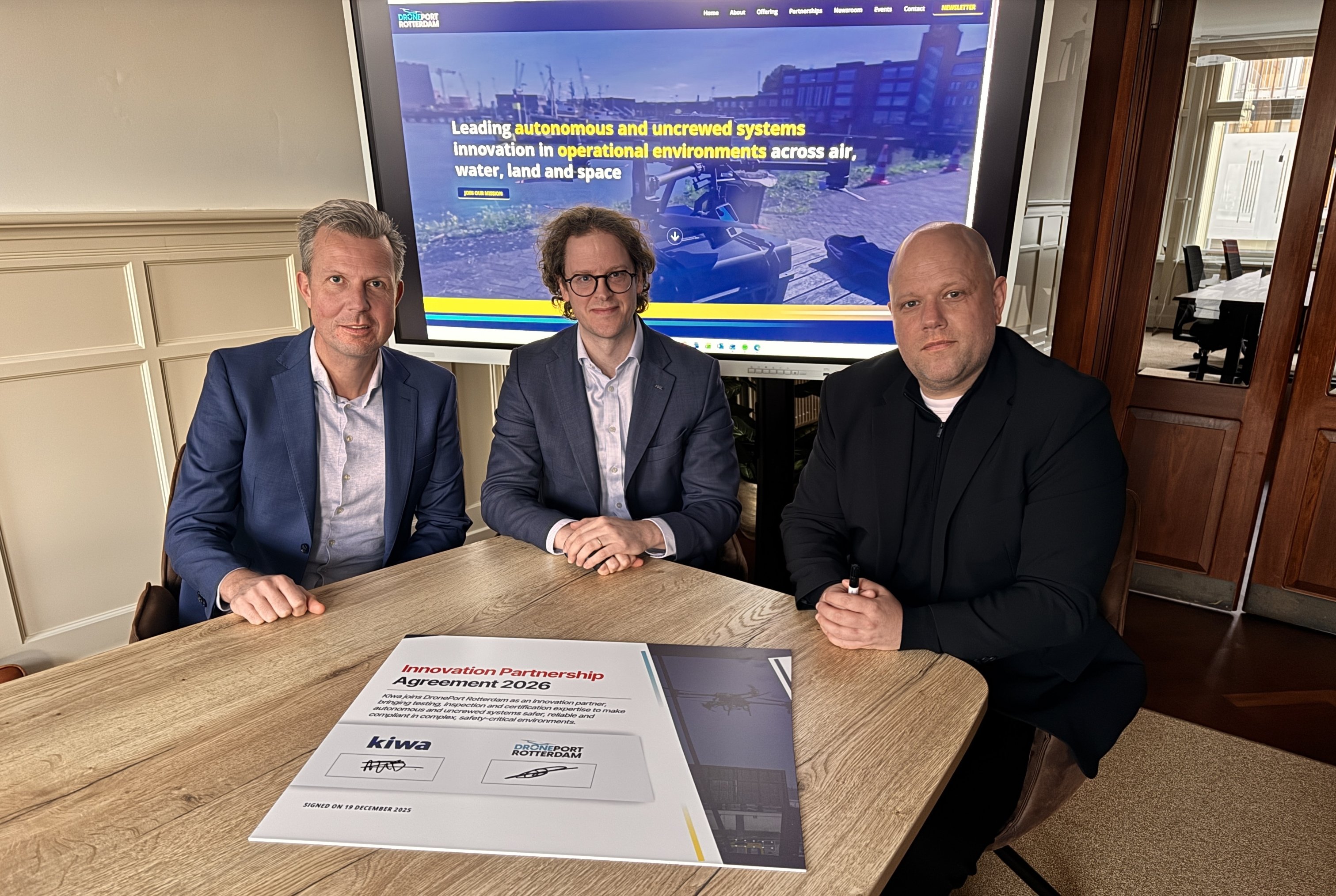Three individuals seated at a table with a document featuring logos, in front of a screen displaying a graphic and text about autonomous and unmanned systems innovation in operational environments.