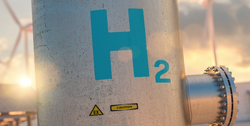 A close-up of a metallic pipe labeled "H2" with a caution sign, set against a backdrop of wind turbines and a sunset.