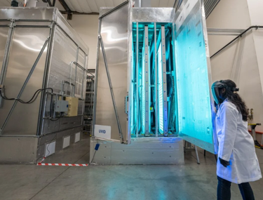 A Kiwa laboratory employee in protective gear observes a large UVC sterilization chamber emitting blue light inside a laboratory setting