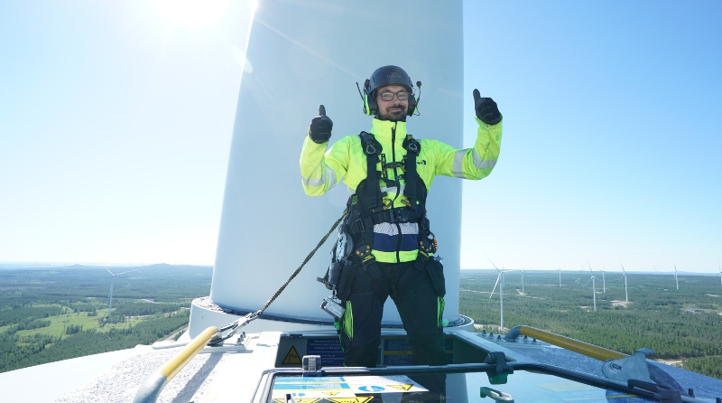 A person in safety gear gives a thumbs-up atop a wind turbine, with a scenic landscape and other turbines in the background.