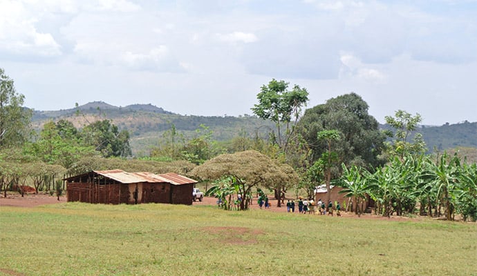 Rural landskap med enkla byggnader, träd och en grupp människor som samlas vid sidan av ett öppet fält under en klar himmel.