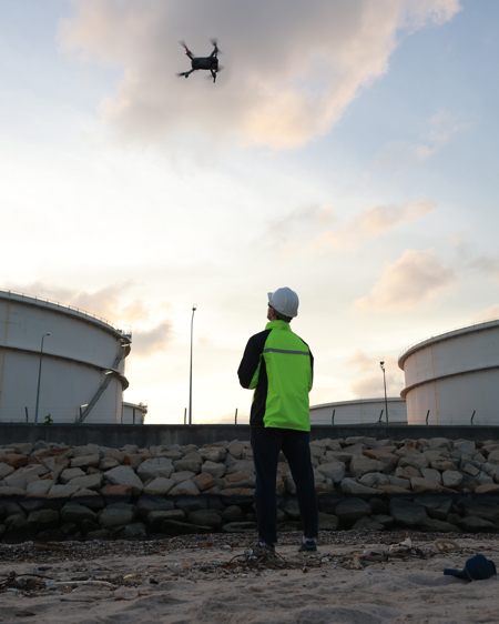 A Kiwa drone pilot during a drone inspection, in a high-visibility jacket and helmet operating a drone near large industrial storage tanks at sunset