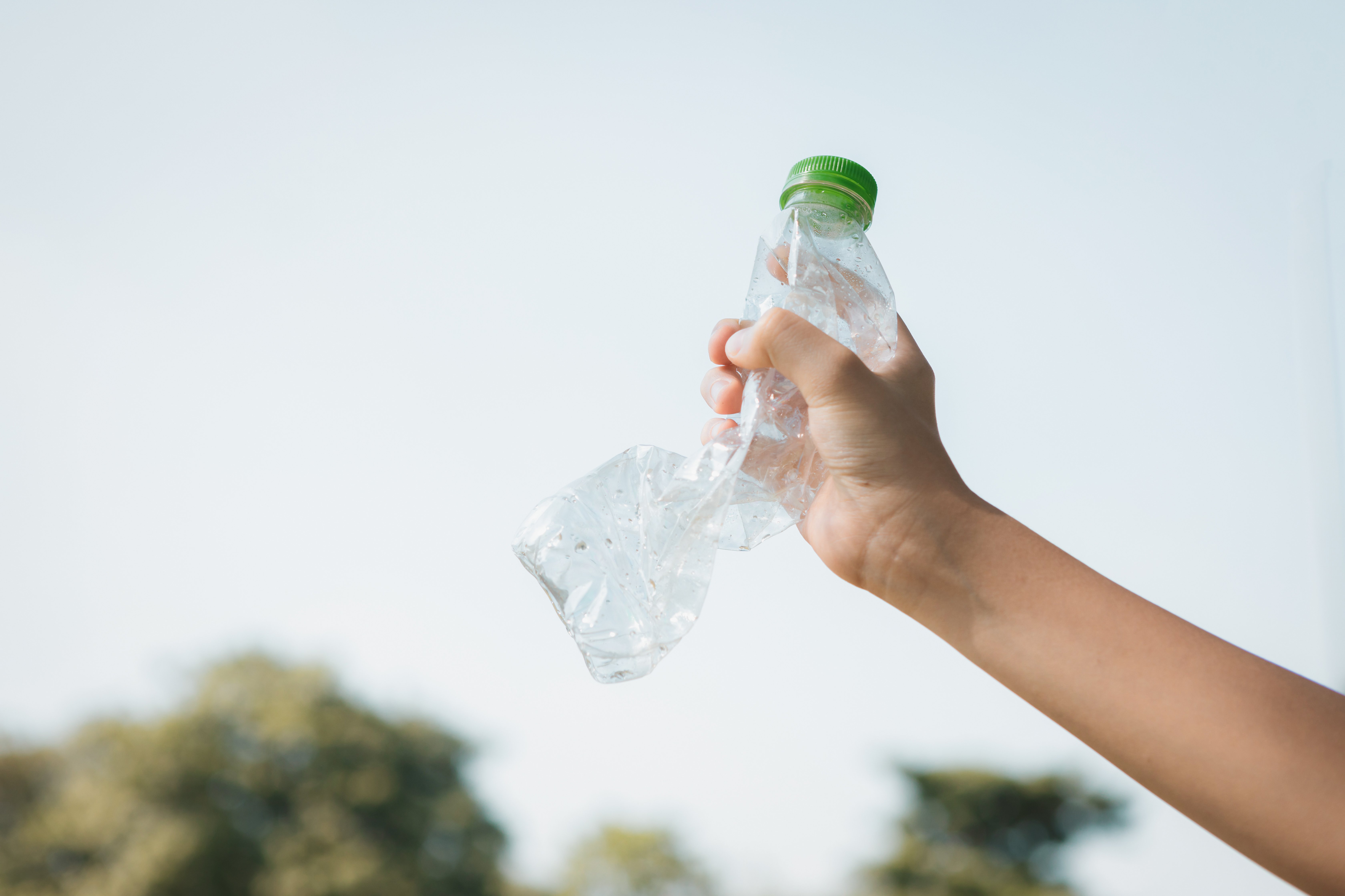 A hand holding a crumpled plastic water bottle against a bright sky background.