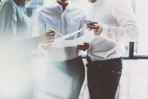 Three business professionals collaborating, examining documents and using a smartphone in a brightly lit office setting