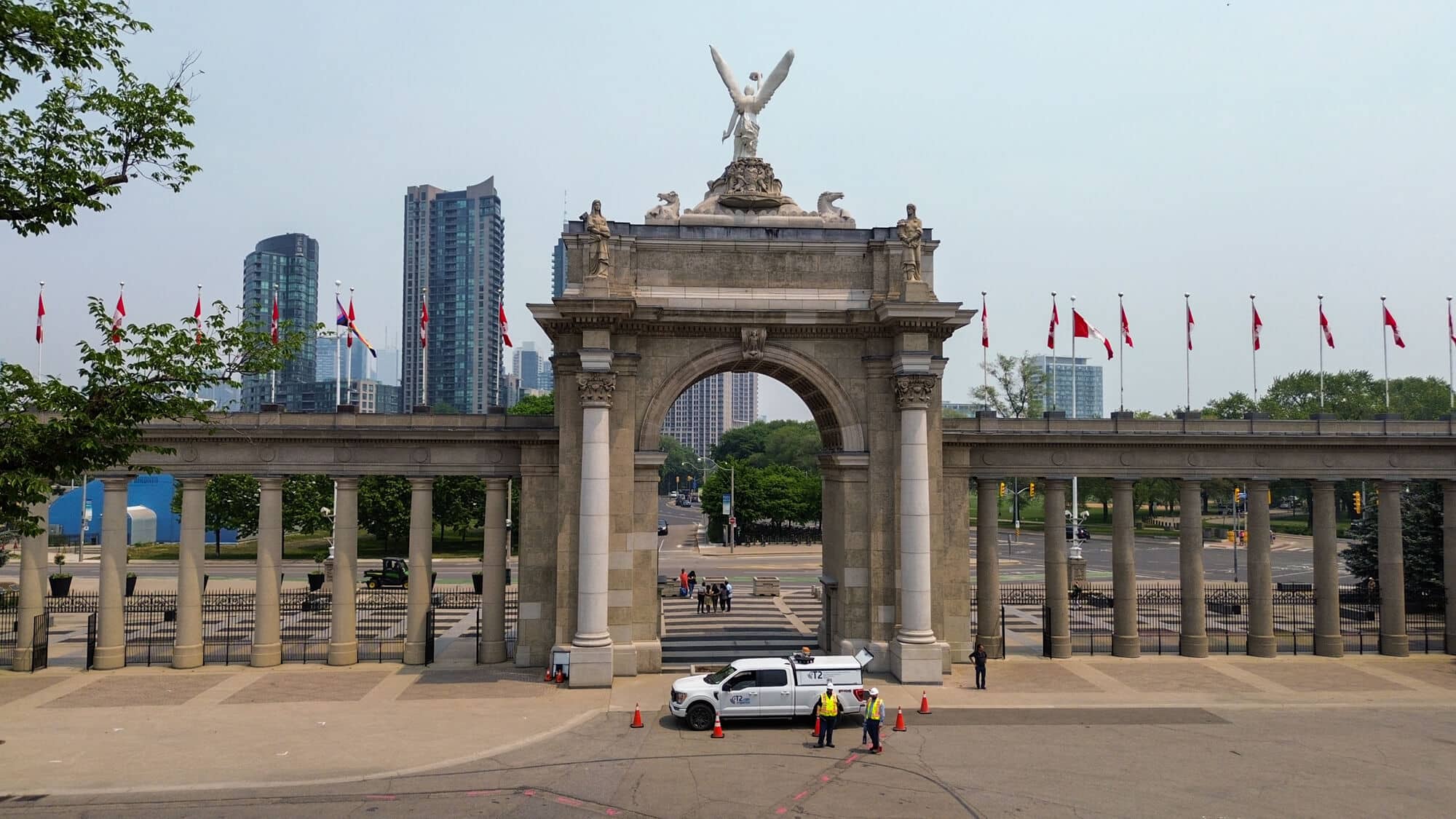 Historic stone archway entrance with Canadian flags, high-rise buildings, and a white Kiwa T2 utility truck in the foreground.