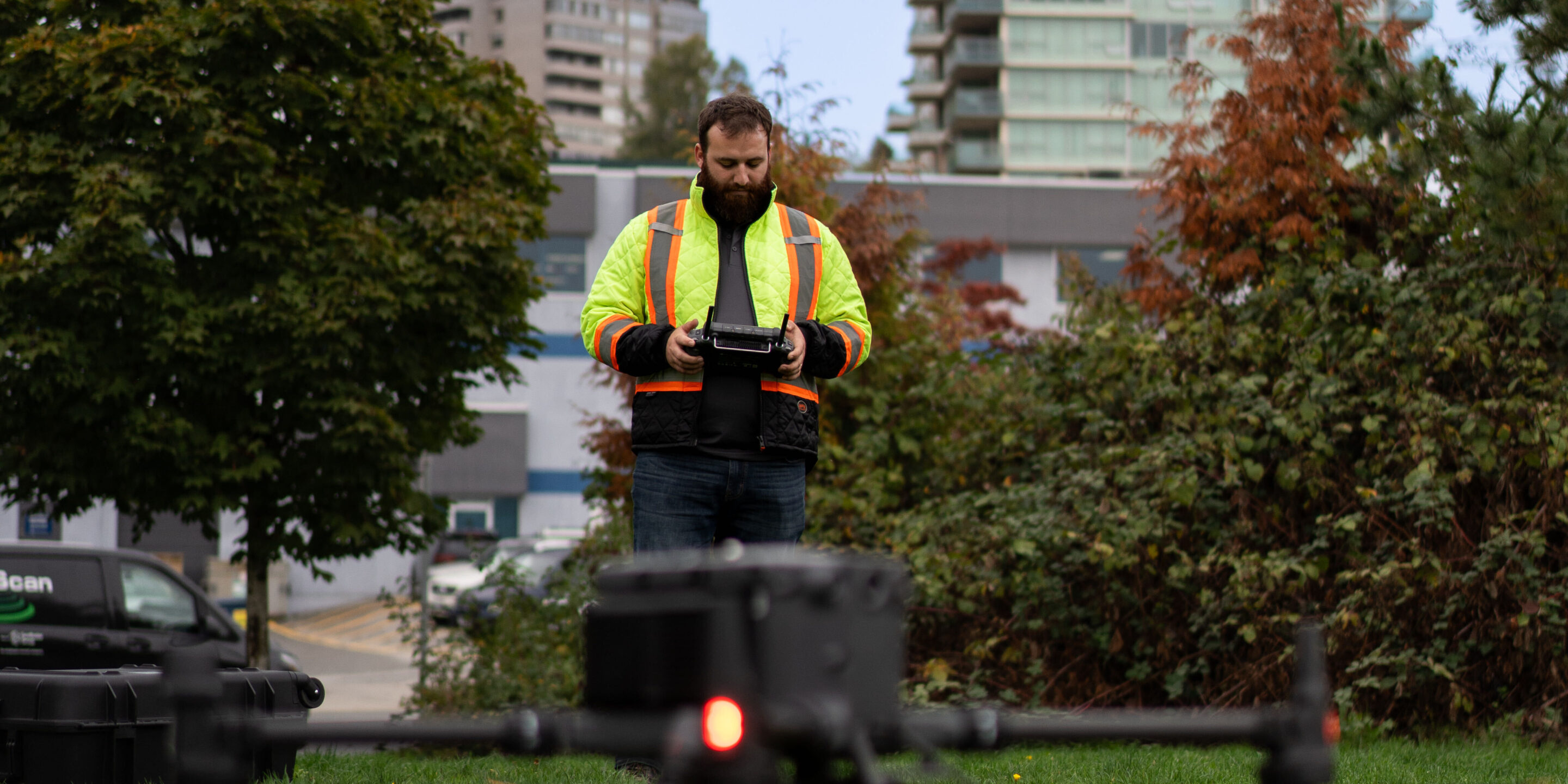 Angus Botting, Kiwa T2 drone operator, operating a drone controller outdoors in an urban park, with city buildings in the background.