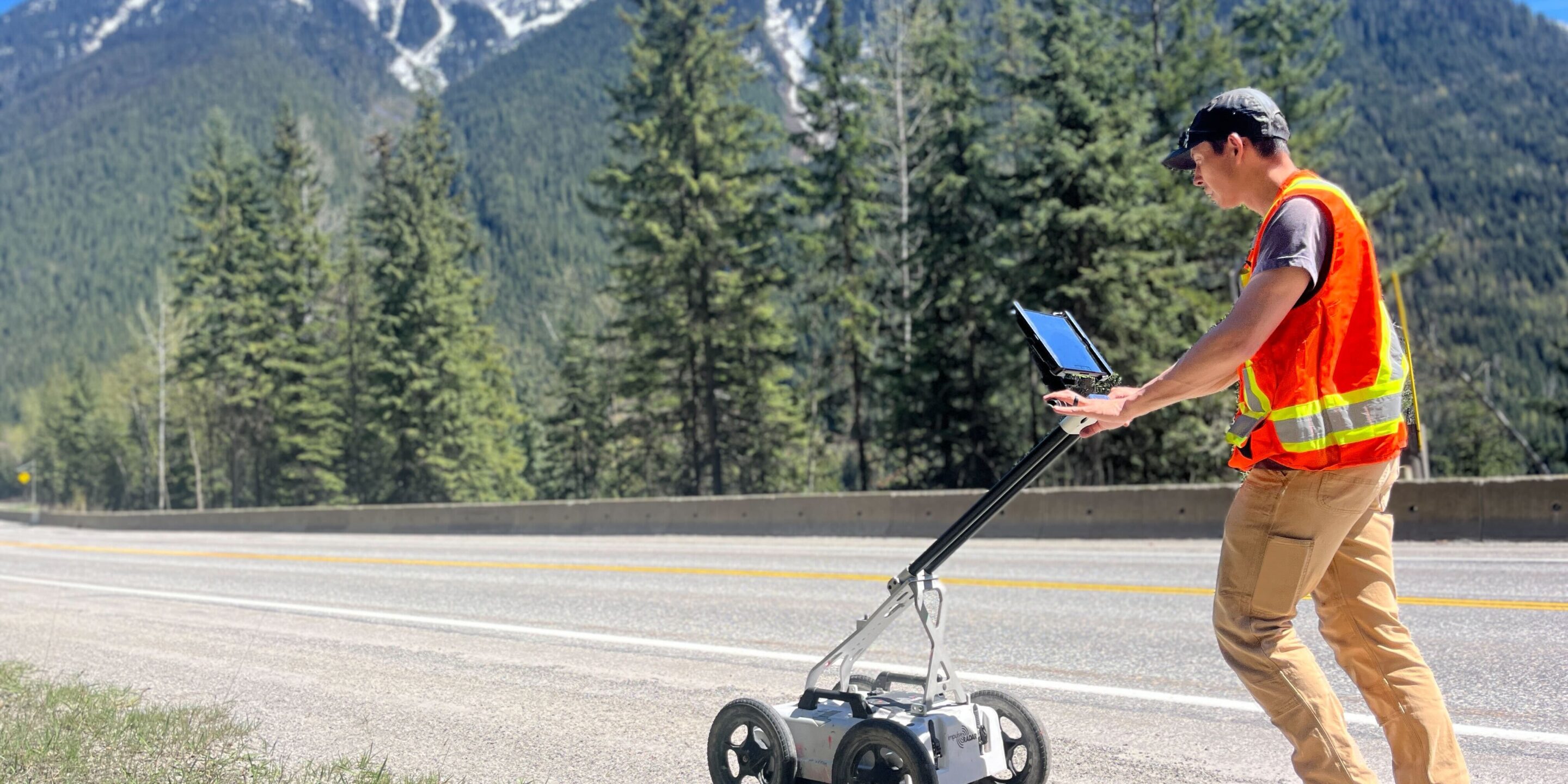 Micheal, a Kiwa T2 survey worker, using a tablet to control a wheeled inspection device on a highway, with forested mountains in the background.