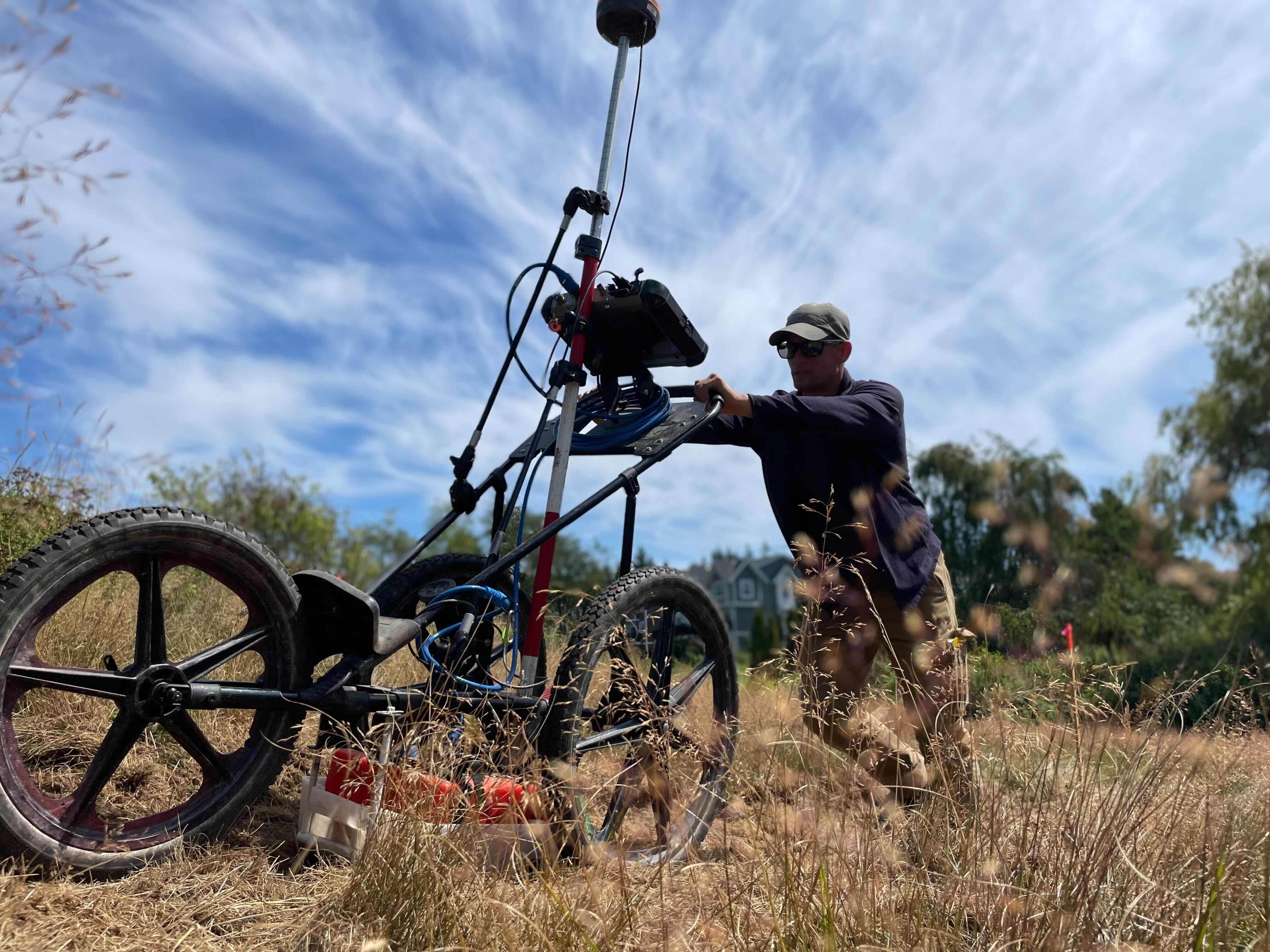 Kiwa T2 employee pushing a wheeled field survey rig in tall grass under a partly cloudy sky, with a mounted sensor and cables.