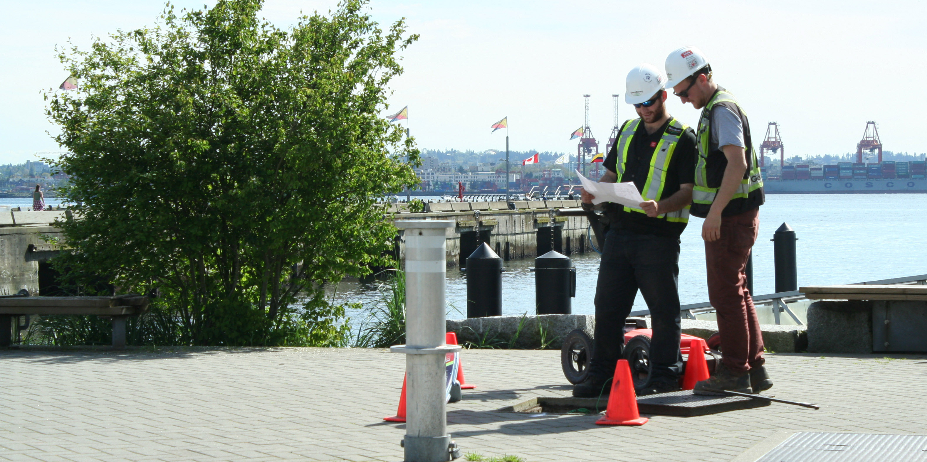 Two Kiwa T2 employees in safety vests review a map near a waterfront dock, with traffic cones and port cranes in the background.