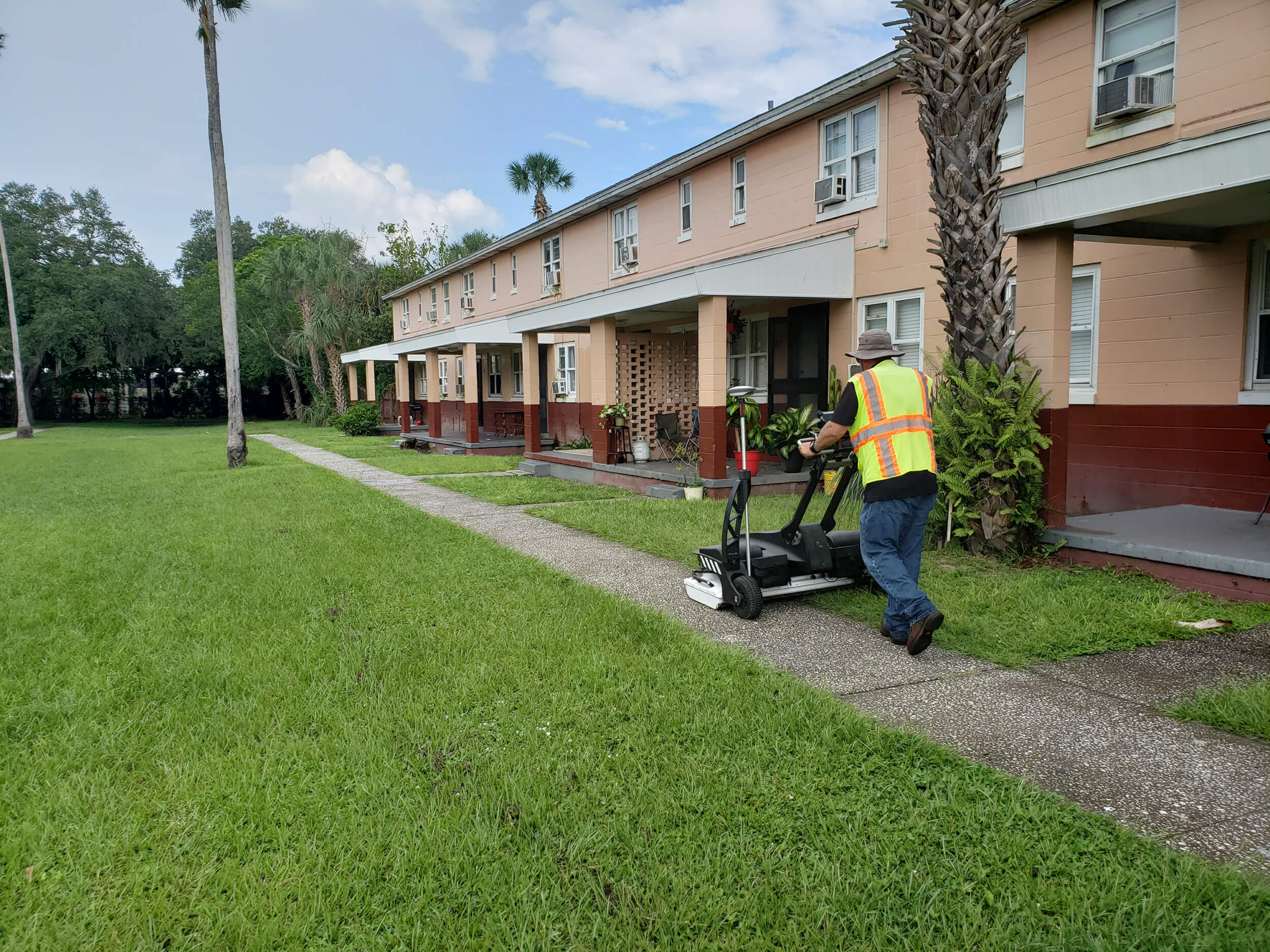 Kiwa T2 employee in a safety vest pushes a lawn mower along a sidewalk beside a row of apartment buildings and palm trees.