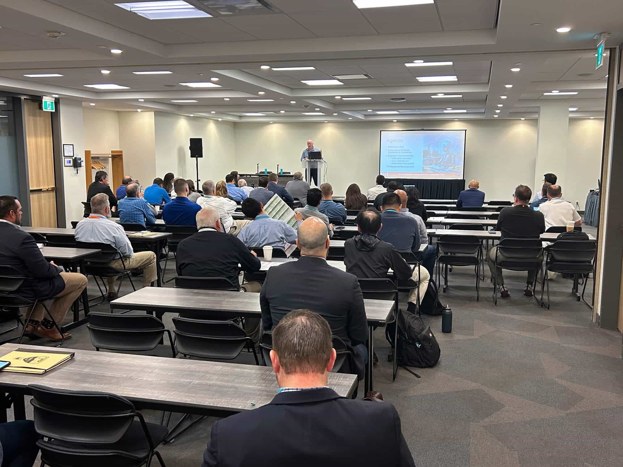 Conference presentation in a meeting room with seated attendees watching a speaker at a podium beside a projection screen.