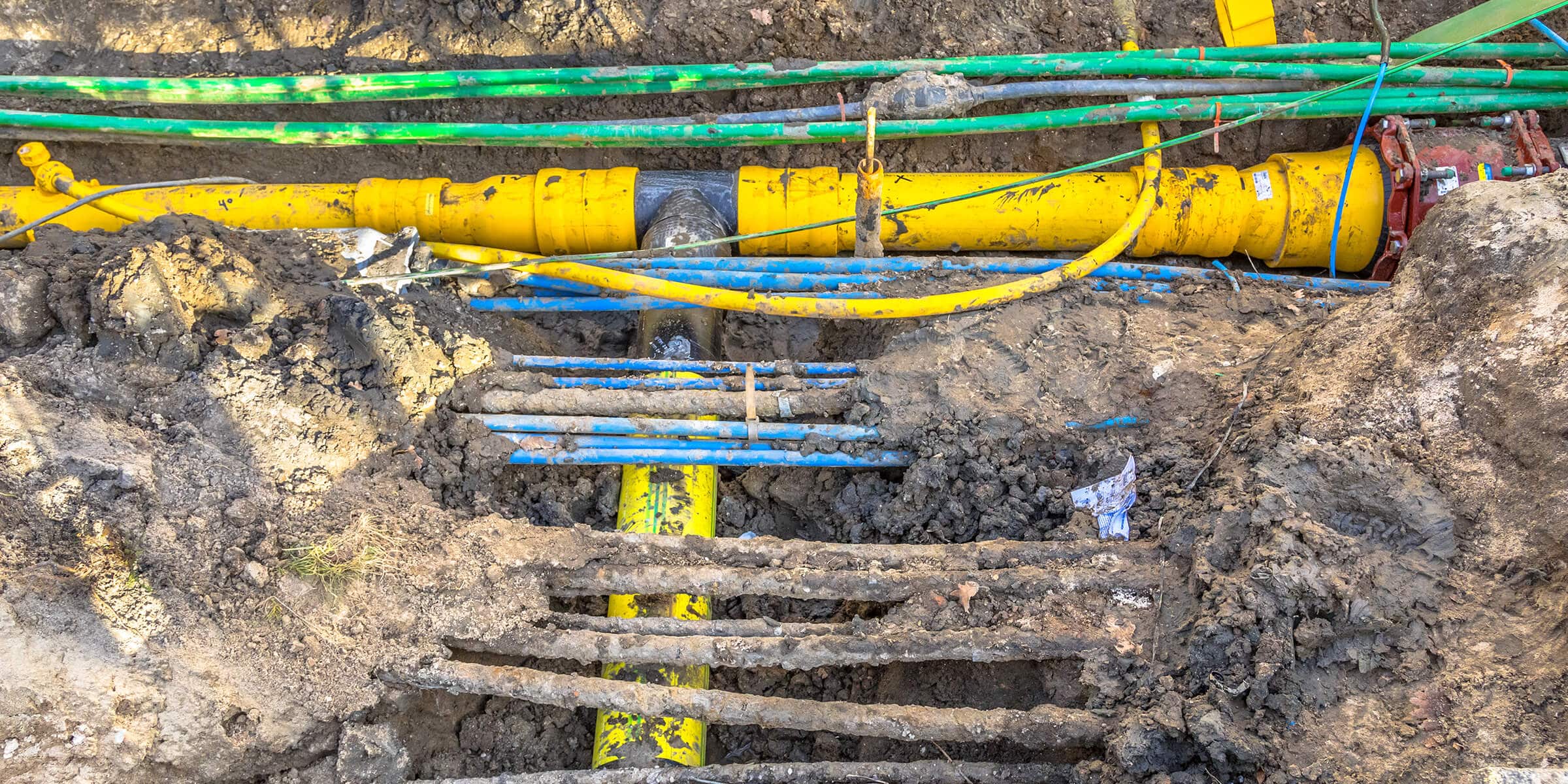 Conduite jaune enterrée dans une tranchée de chantier, avec tuyaux et câbles visibles dans la boue.