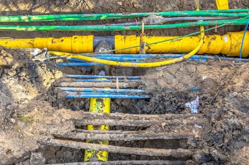 Conduite jaune enterrée dans une tranchée de chantier, avec tuyaux et câbles visibles dans la boue.