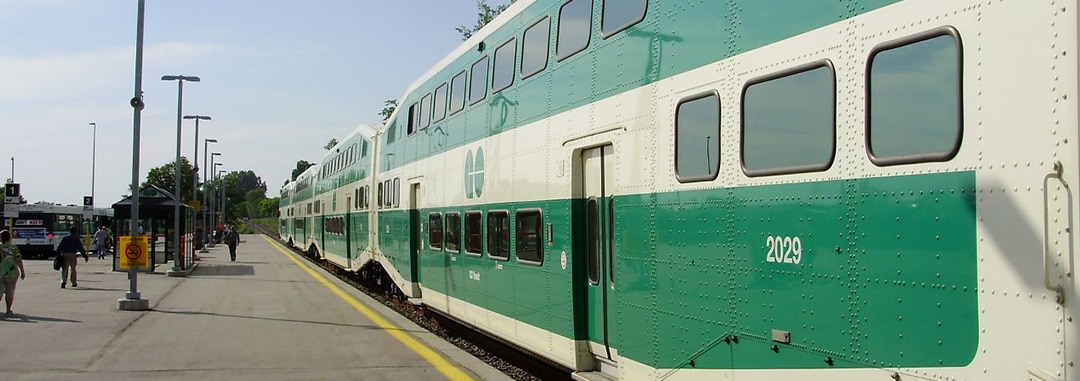 Green and white double-decker passenger train at a station platform, with people and platform shelters in the background.
