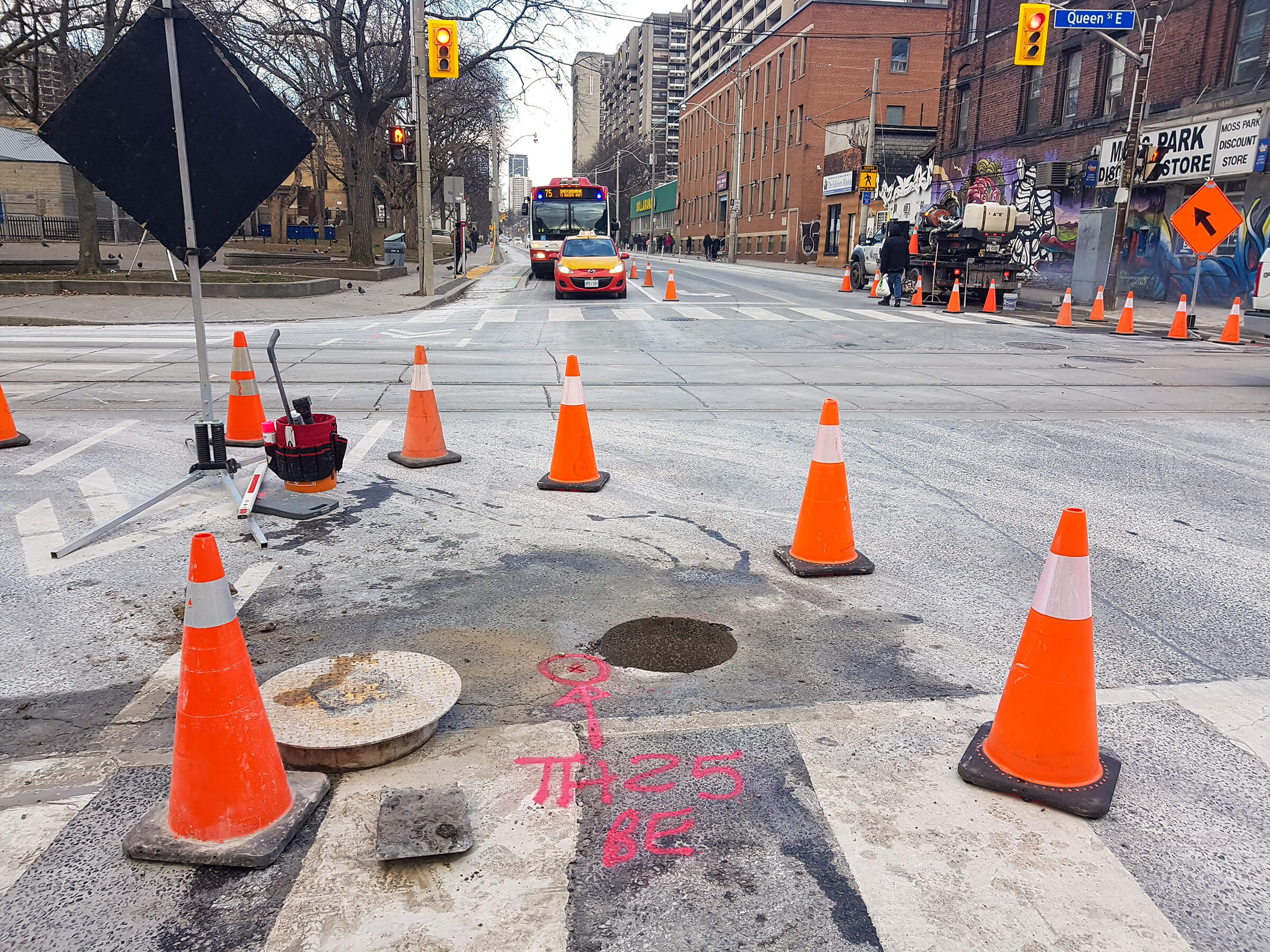 Traffic cones and roadwork markers on a city street intersection with a bus, taxi, and Queen Street East sign in the background.