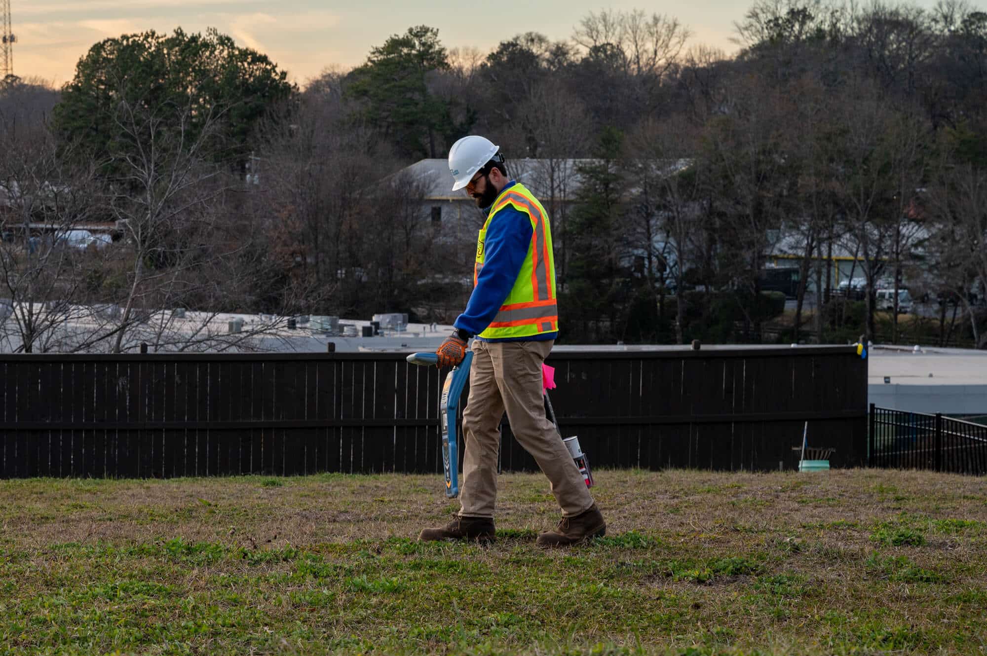 Kiwa T2 employee in a hard hat and high-visibility vest walking on a grassy site at dusk, carrying a blue tool or measuring board.