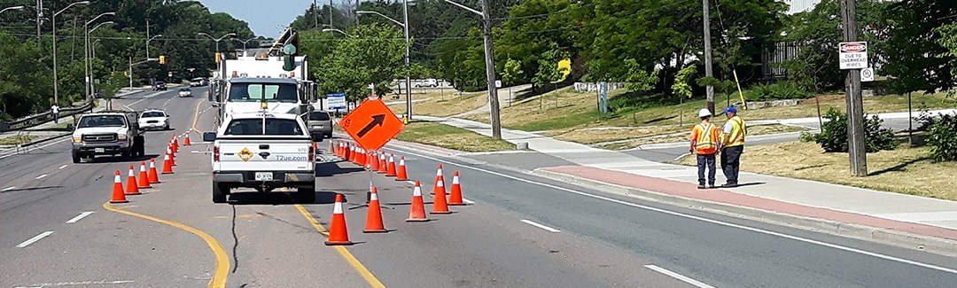 Roadwork scene with orange traffic cones, utility trucks, and two Kiwa T2 workers standing beside a closed lane on a suburban street.