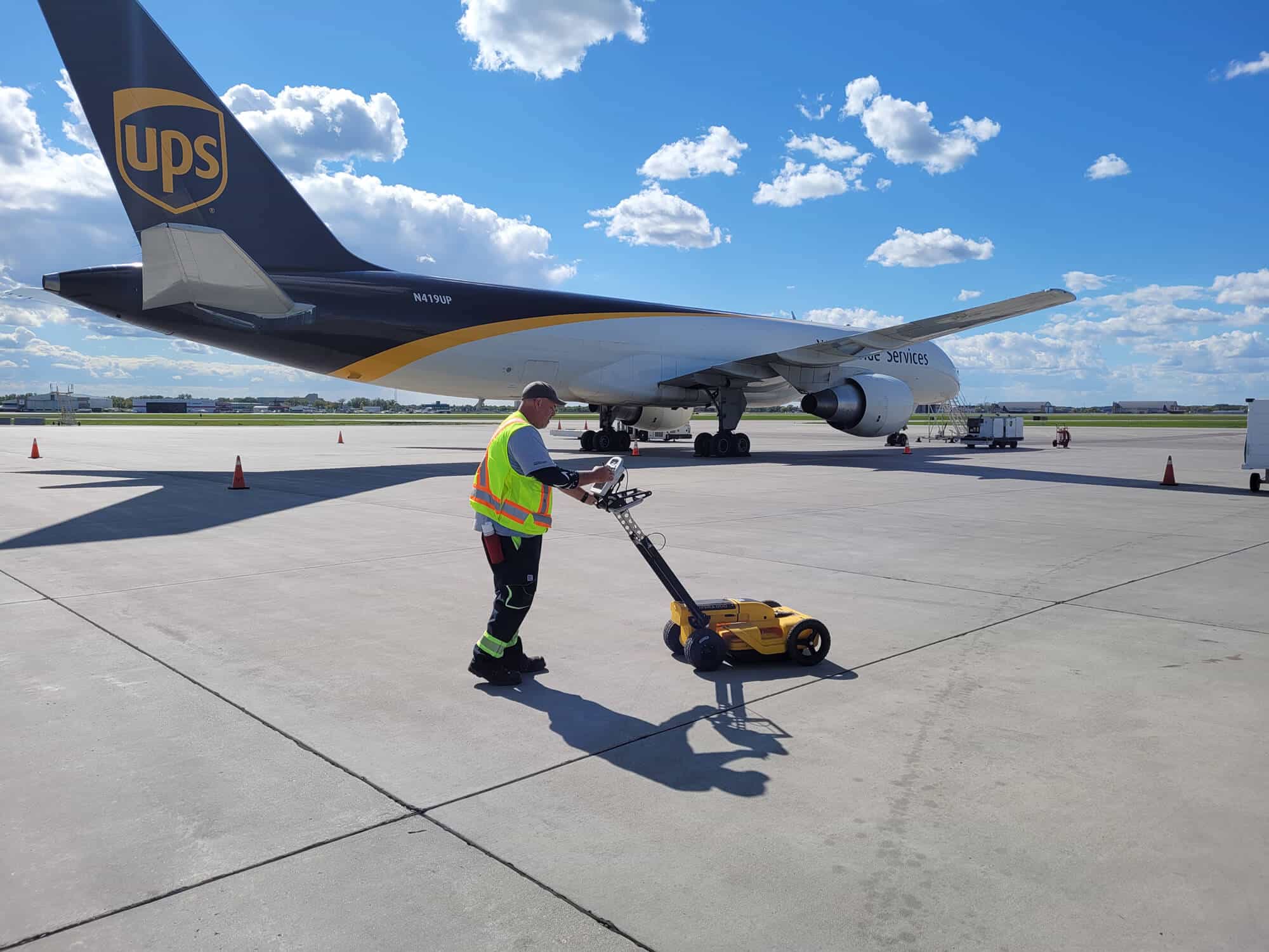 Kiwa T2 employee operating a yellow line-marking machine near a UPS cargo airplane on the tarmac under a blue sky.