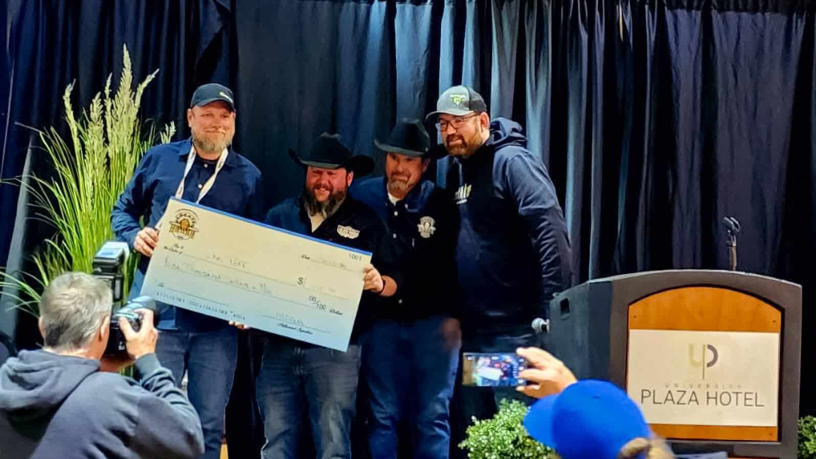 Four men pose with a large ceremonial cheque on a hotel stage during a charity presentation event.