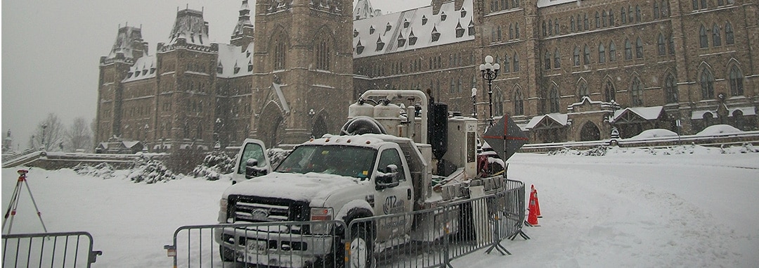 Snow-covered Kiwa T2 utility truck parked in front of a large Gothic-style government building (Ottowa parliament) during winter snowfall.