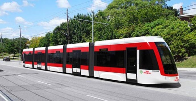 Modern red and white light rail train travelling on a street beside green trees under a blue sky.