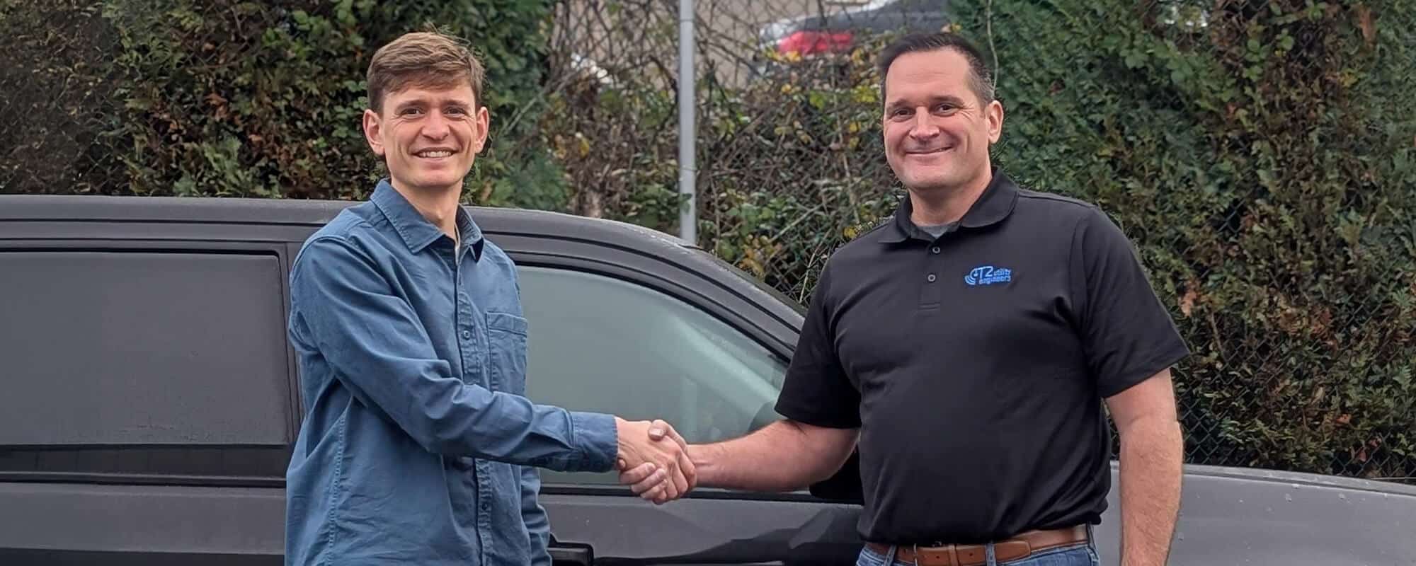 Two men shaking hands beside a black vehicle outdoors, with greenery in the background.