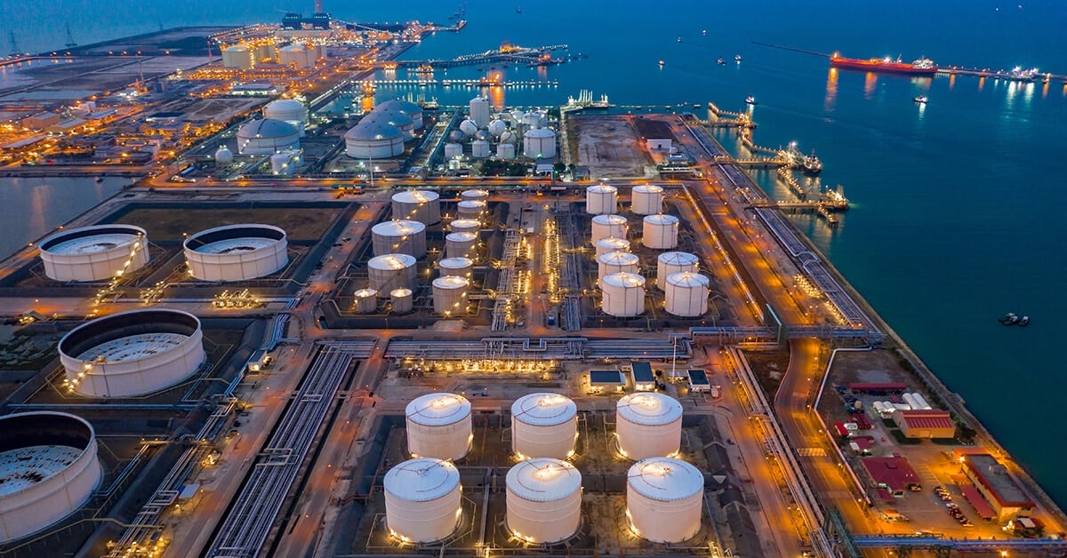 Aerial view of an illuminated oil refinery and storage tanks at a waterfront industrial port at dusk.