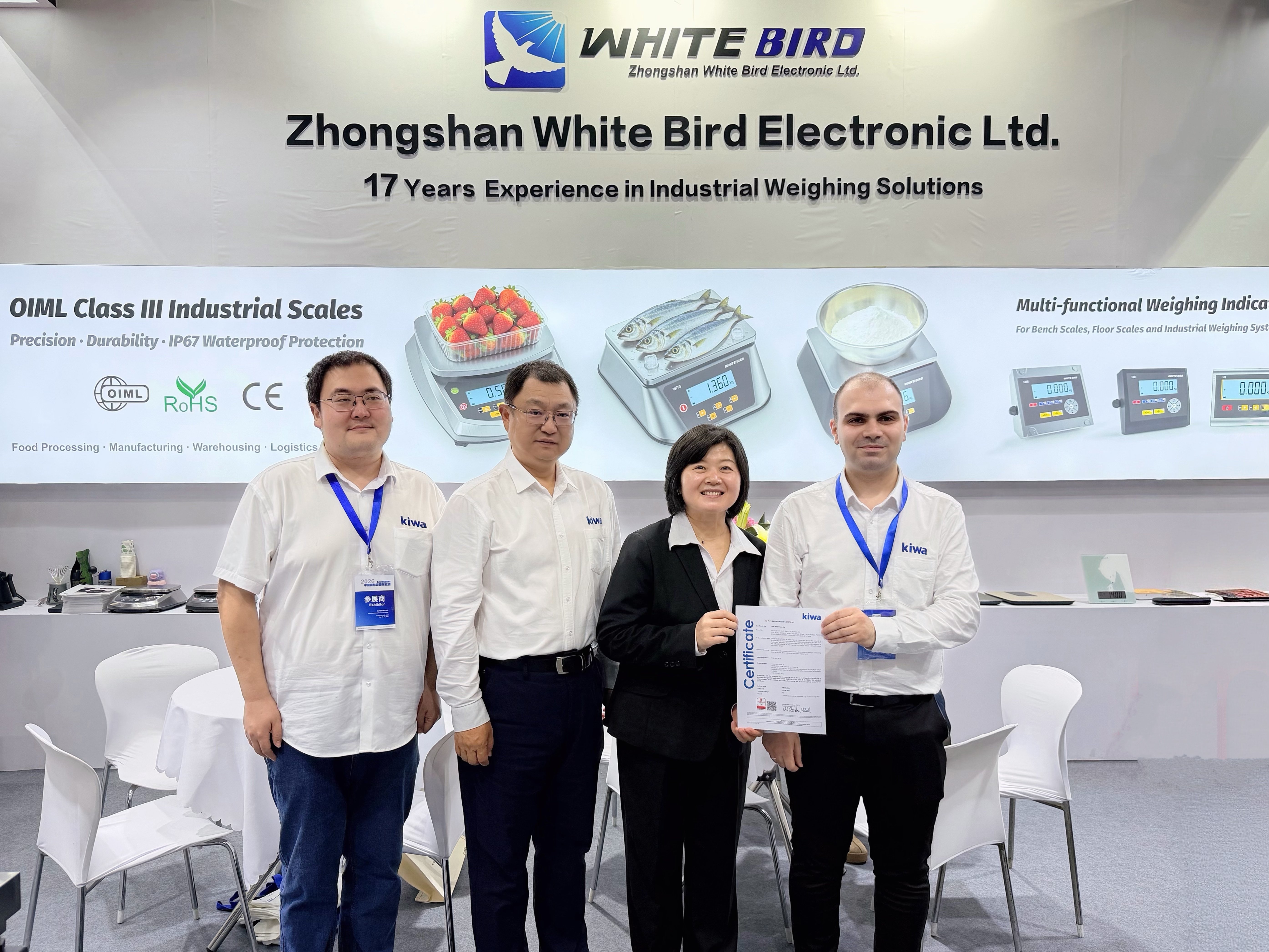 在工业衡器展台前的四人合影，一人手持证书，背景展示 Zhongshan White Bird Electronic Ltd. 的电子秤产品宣传板。