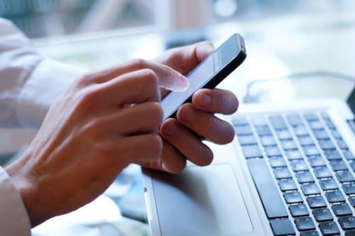 Två händer håller i en mobil, som används över en bärbar dators tangentbord.