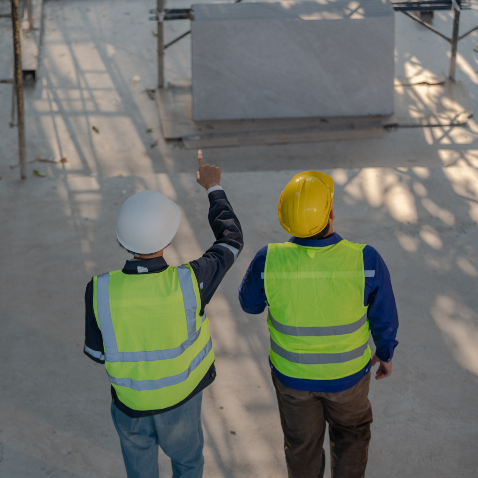 Dos trabajadores de construcción con cascos y chalecos reflectantes inspeccionan una obra; uno señala hacia arriba mientras ambos caminan sobre una superficie de concreto.