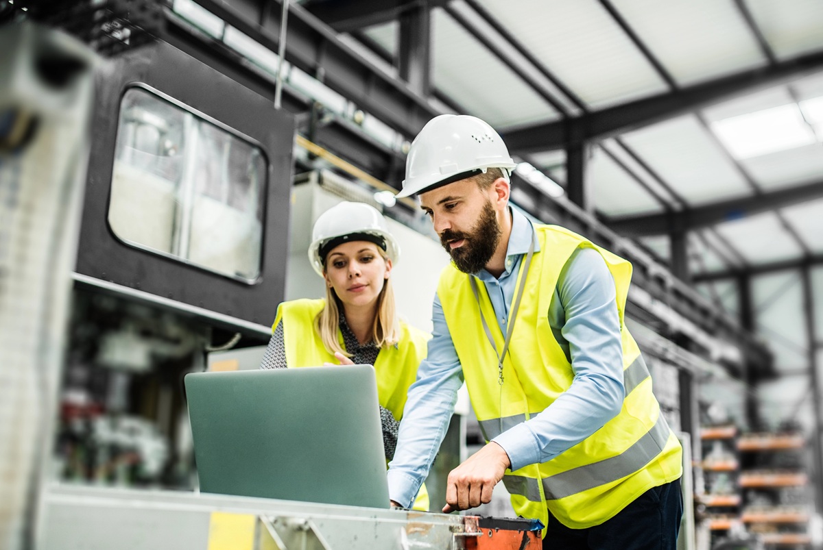 Two individuals in a factory setting wearing white safety helmets and yellow high-visibility vests, one using a laptop and the other standing beside. Industrial equipment and shelves are visible in the background.