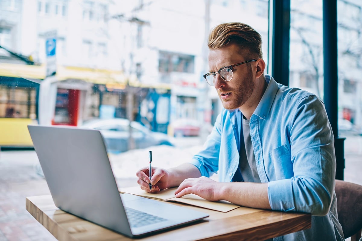 A person sitting at a wooden table in a cafe, writing on a notepad with a pen, with an open laptop and large windows showing an urban street view in the background.