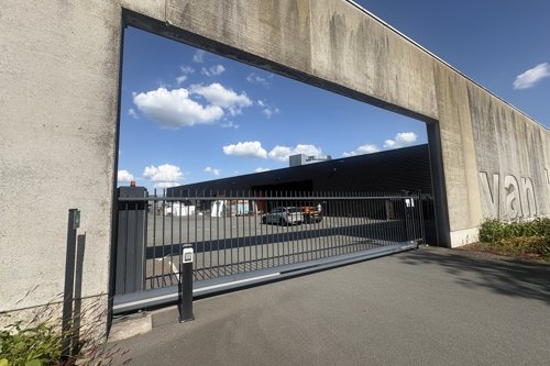 Moderne industriële ingang met een grote betonnen poort, metalen schuifhek en een afgesloten terrein onder een blauwe lucht met wolken.