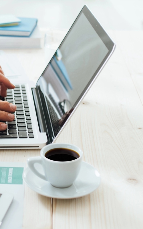 Man typing on a laptop at a wooden desk with coffee cup, tablet, and documents nearby.